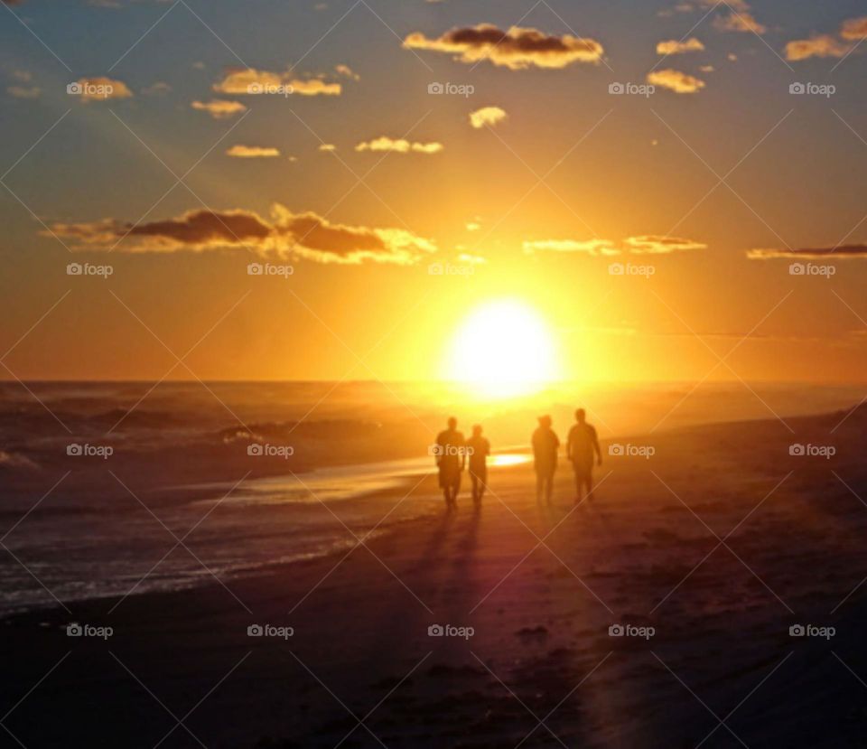 Couples walk along the sandy beach during sunset. The golden disc is already touching the surface of the ocean. It seems in a hurry to disappear, slipping quickly behind the line of the horizon spreading its last rays