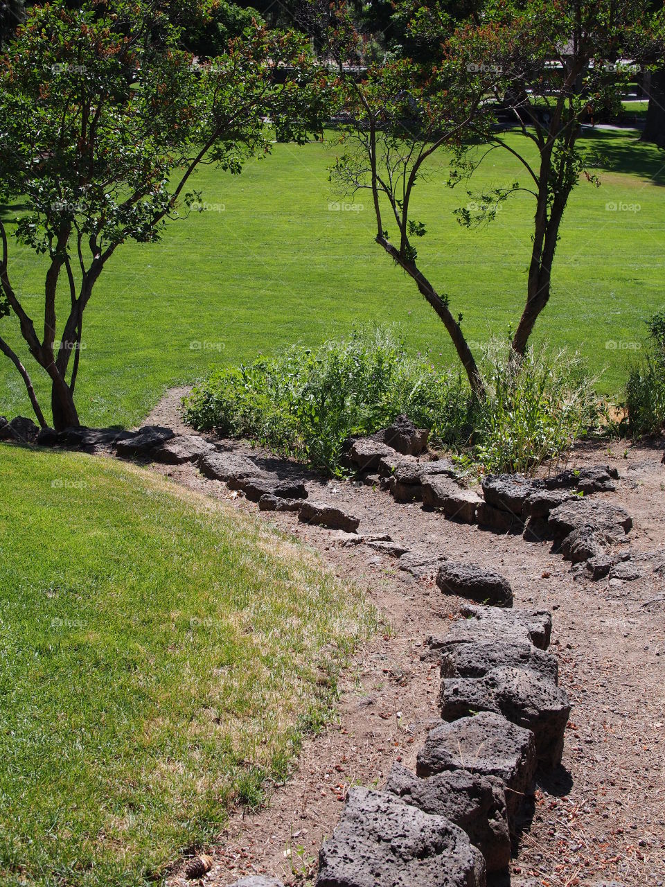 A dirt path leads through tiers with rock walls of different growth from grass to flowers and trees in Pioneer Park in Bend in Central Oregon on a beautiful sunny summer day. 