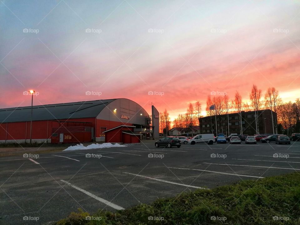 The evening sun setting behind the big hall. Cars in the parking lot.
