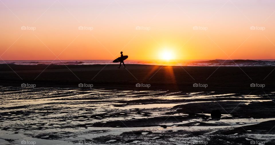 A surfer is silhouetted by the sunset as they walk across the beach at Pataias 