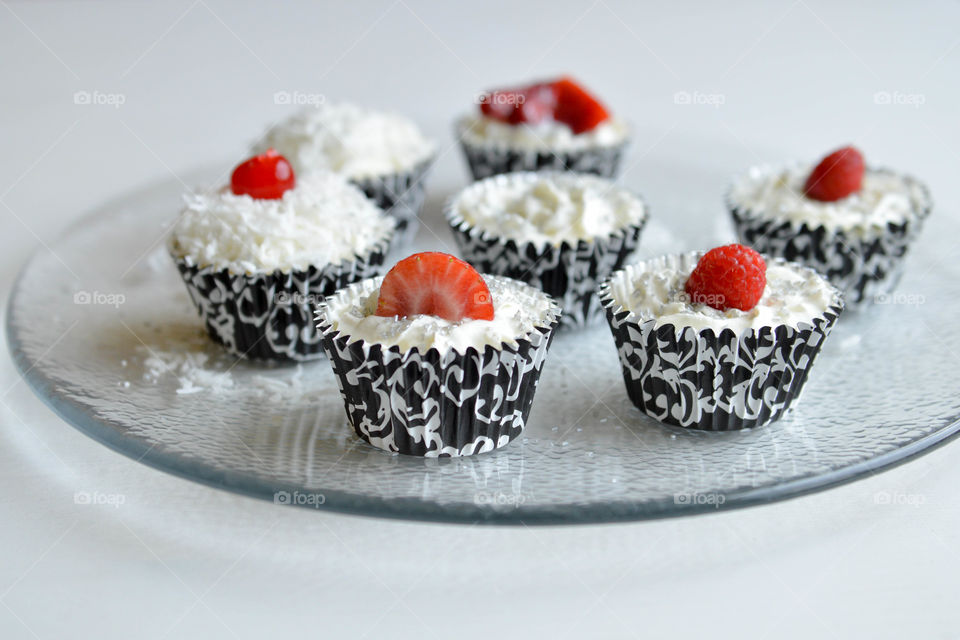 Mini cheesecakes in black and white decorative wrappers topped with whipped cream, coconut shavings and berries