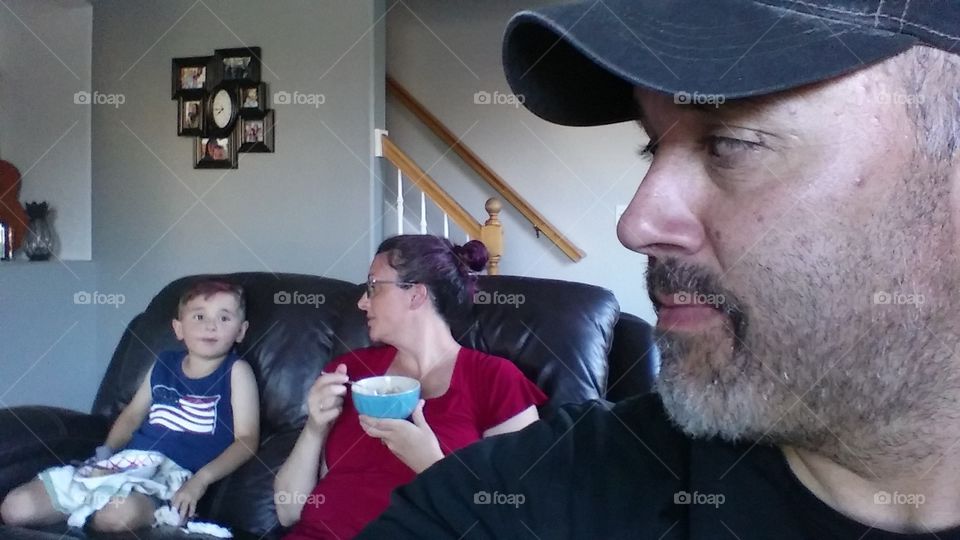 A family during the Fourth of July eating and sitting together. Father looking over his shoulder to his wife and young son, who are sitting on the couch.