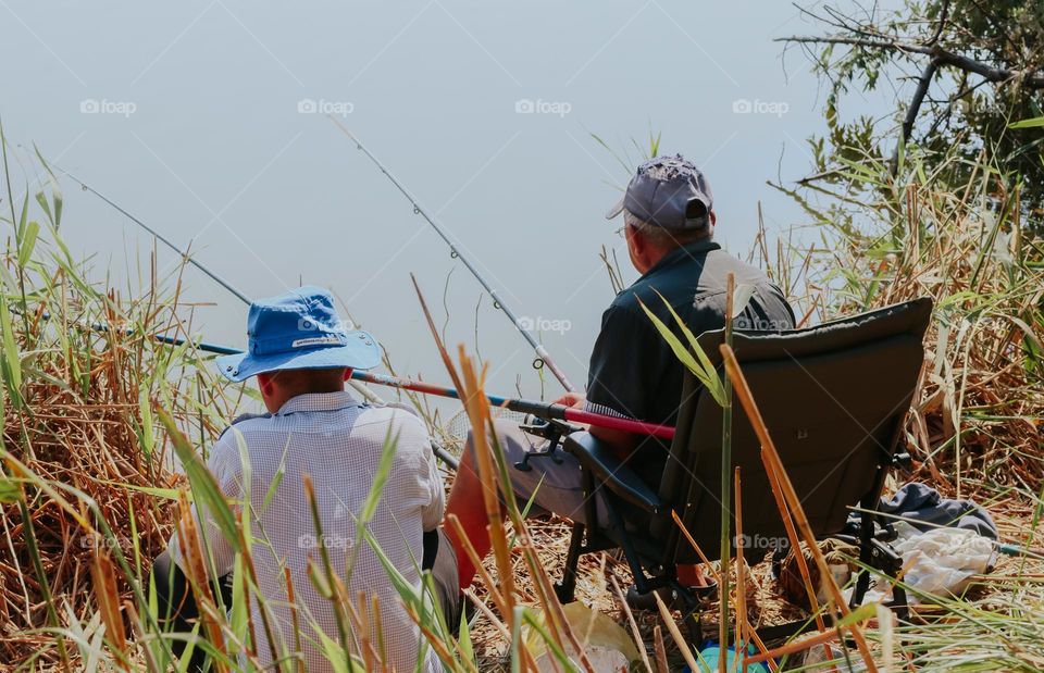 Two Caucasian men wearing panama hats from behind sit in the grass on the river bank and fish on a summer day, close-up side view.