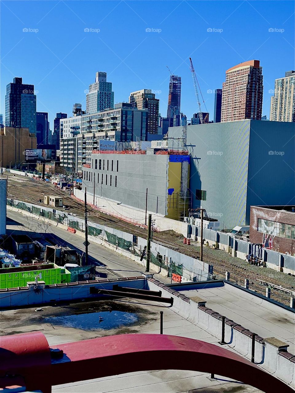 This is the view from the red metal staircase of the “Pulaski Bridge” at “Newtown Creek” in LIC overlooking the “Long Island Railroad” tracks and “iFly”, an indoor flight simulator. In the distance we see “Manhattan”. 2024. Hypnotic Productions