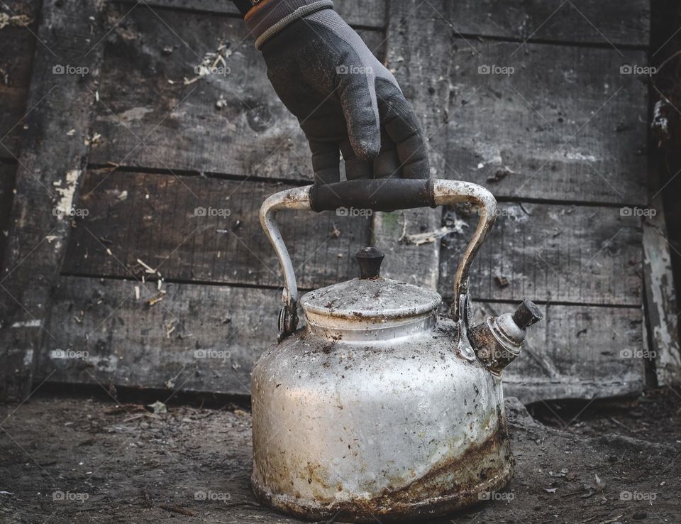 A man's hand in construction gloves holds an old retro aluminum teapot on the ground with a wooden foyer, close-up side view. The concept of retro objects, vintage dishes, metal.