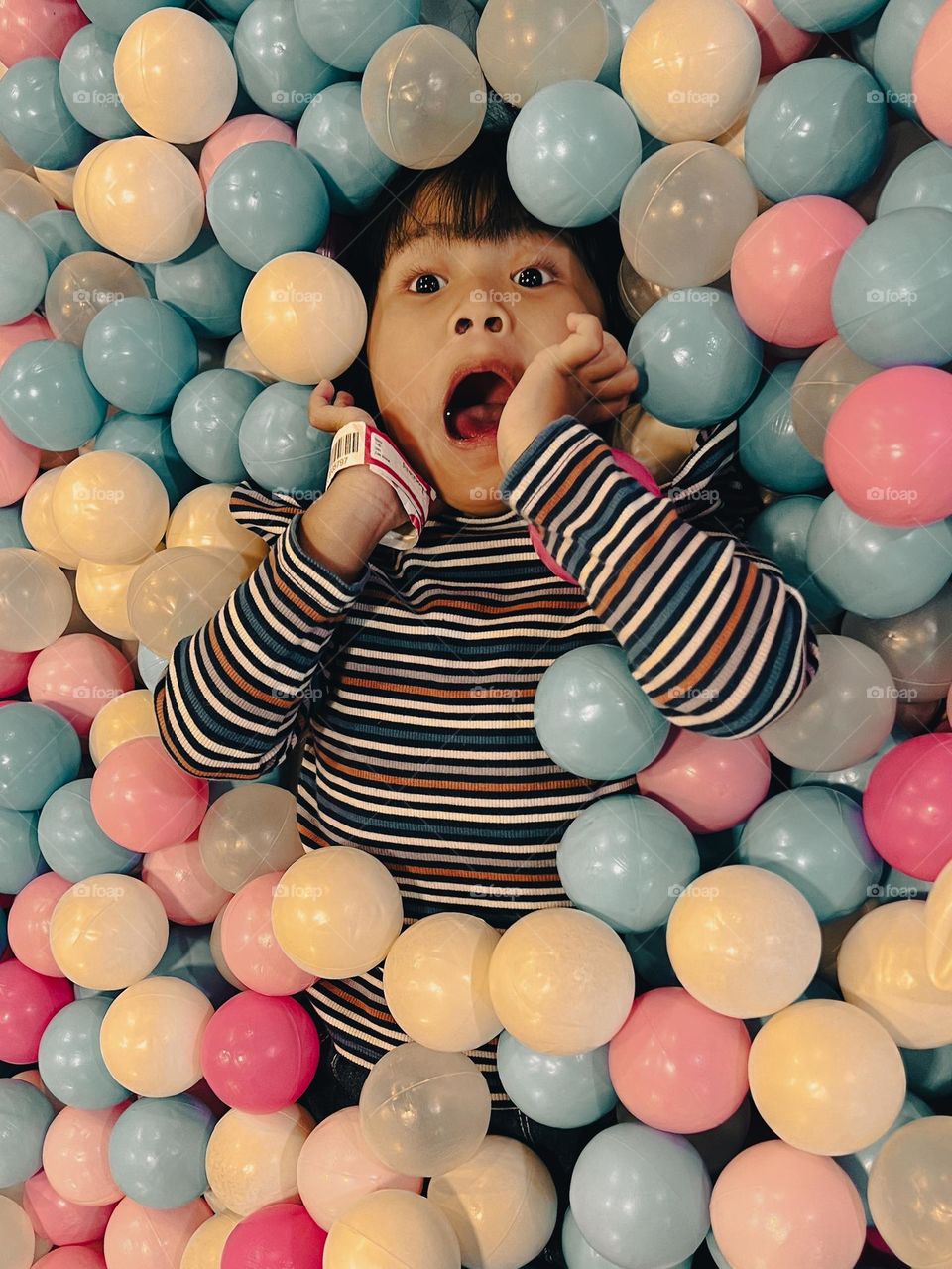 Portrait of a little Asian girl playing in a ball pool playground, lying down with colorful plastic balls in a mall with a shocked expression.