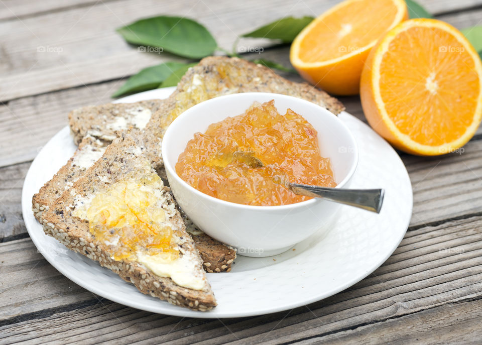 Buttered toast spread with marmalade on  a white plate with a small white bowl filled with marmalade and a spoon. A wooden background and two orange halves compliment the main dish.