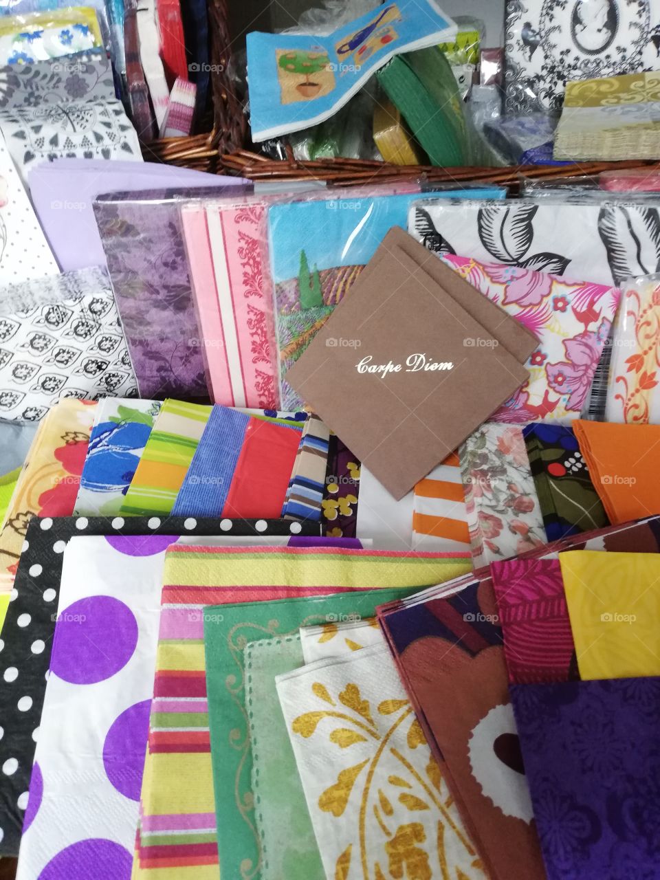 Different napkins in their plastic packages in the brown rattan basket, some are on the table one on the other. Many colours, figures, spots, drawings, texts, in the front carpe diem on the brown napkin