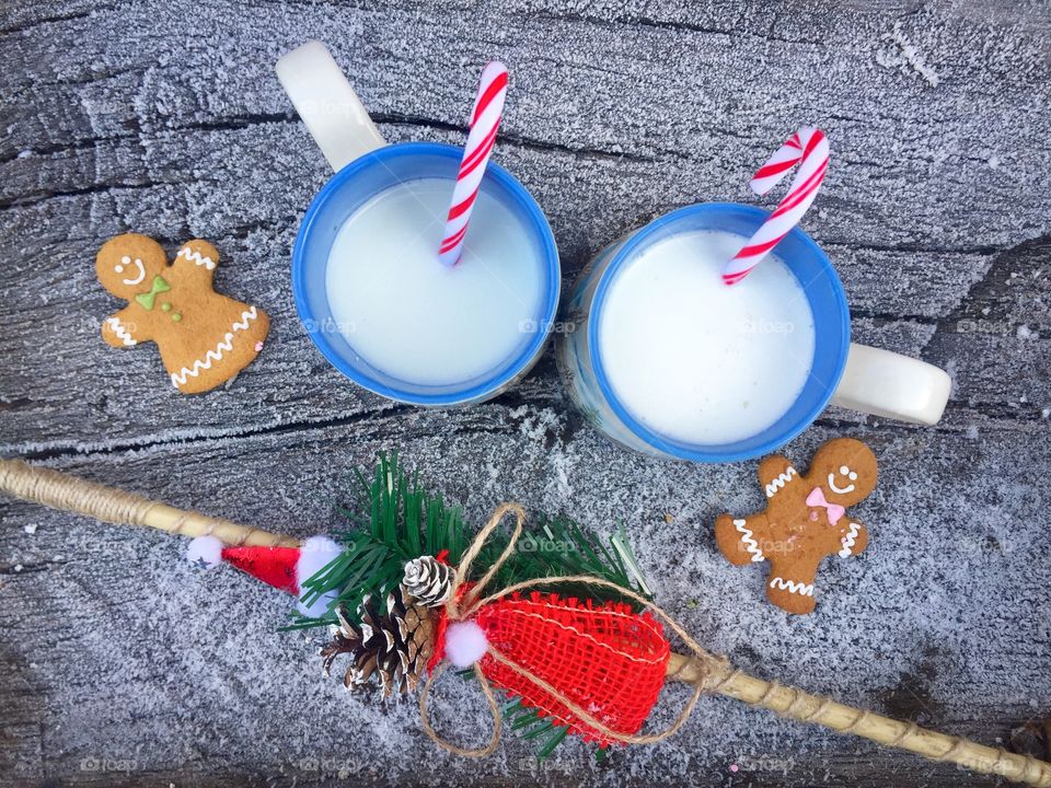Two mugs of white chocolate and milk with candy canes inside on snowy table with two gingerbread men beside