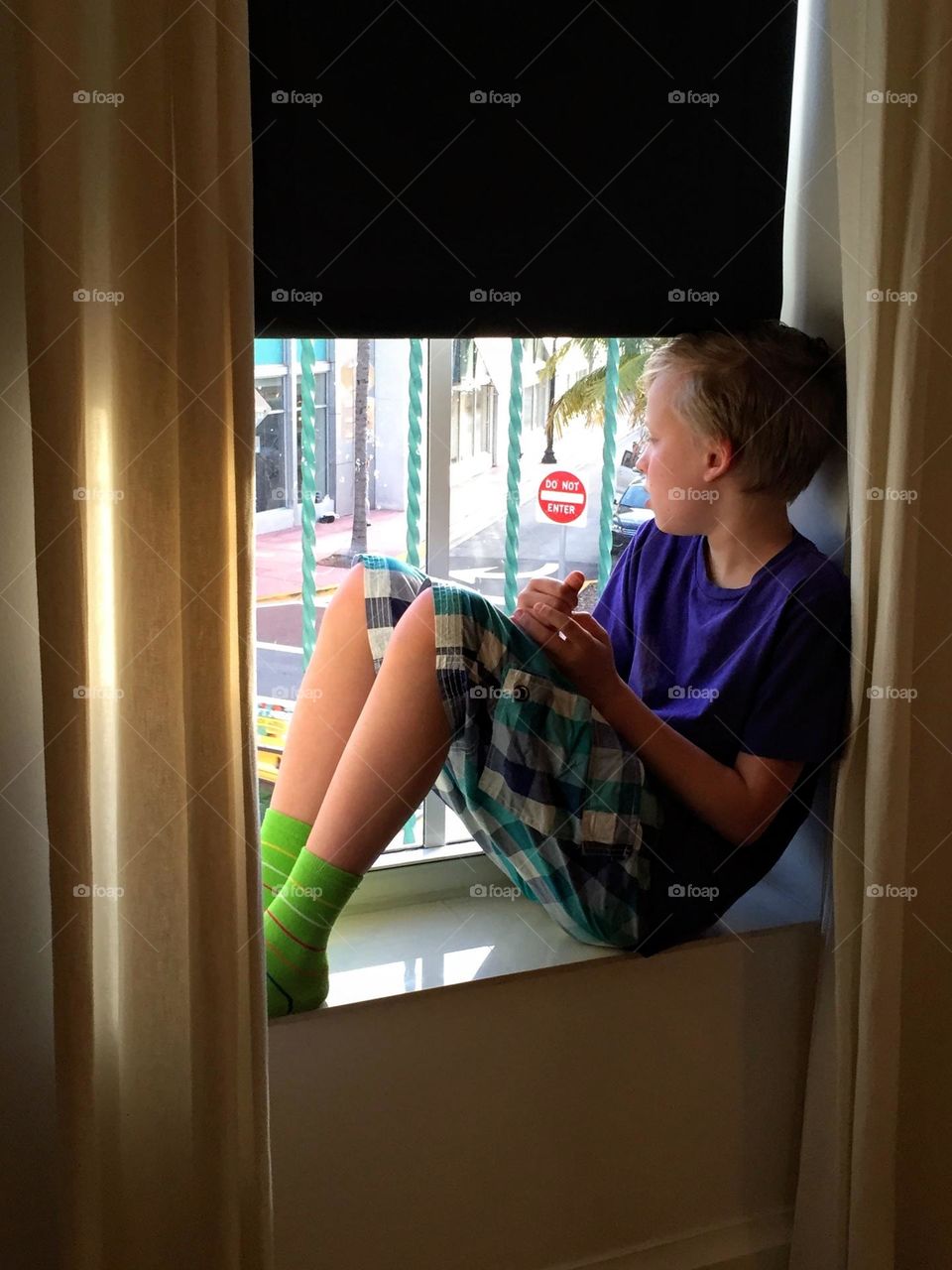 Young boy in checkered shorts sitting on the narrow windowsill and looking out from the window