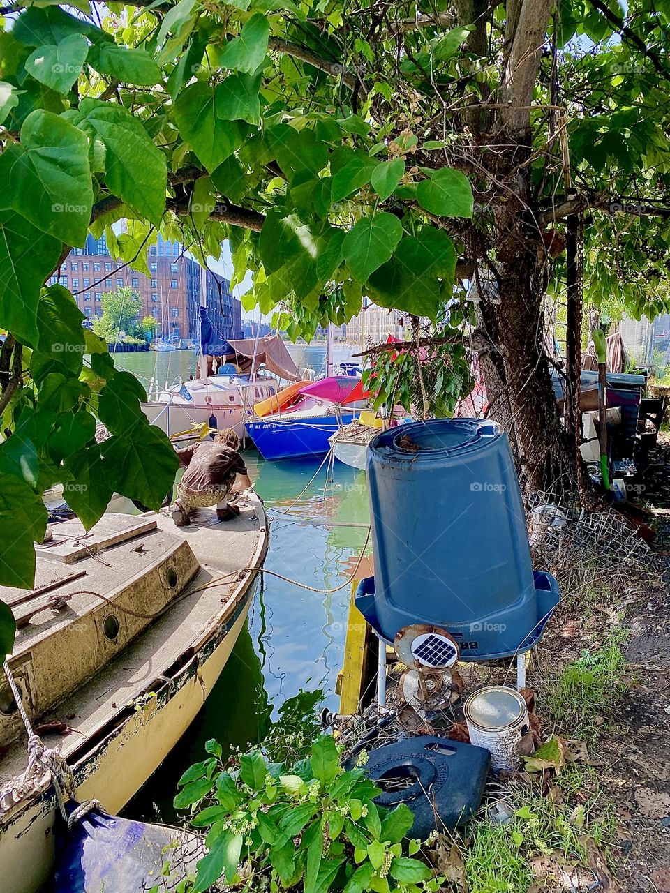 Through the foliage of this tree kneeling on the boat is „Timothy Trespas“ attending to some kind of problem. He is quite good at this sort of thing. We are all tied together here at „Newtown Creek“ in LIC, Queens. 2023. Hypnotic Productions