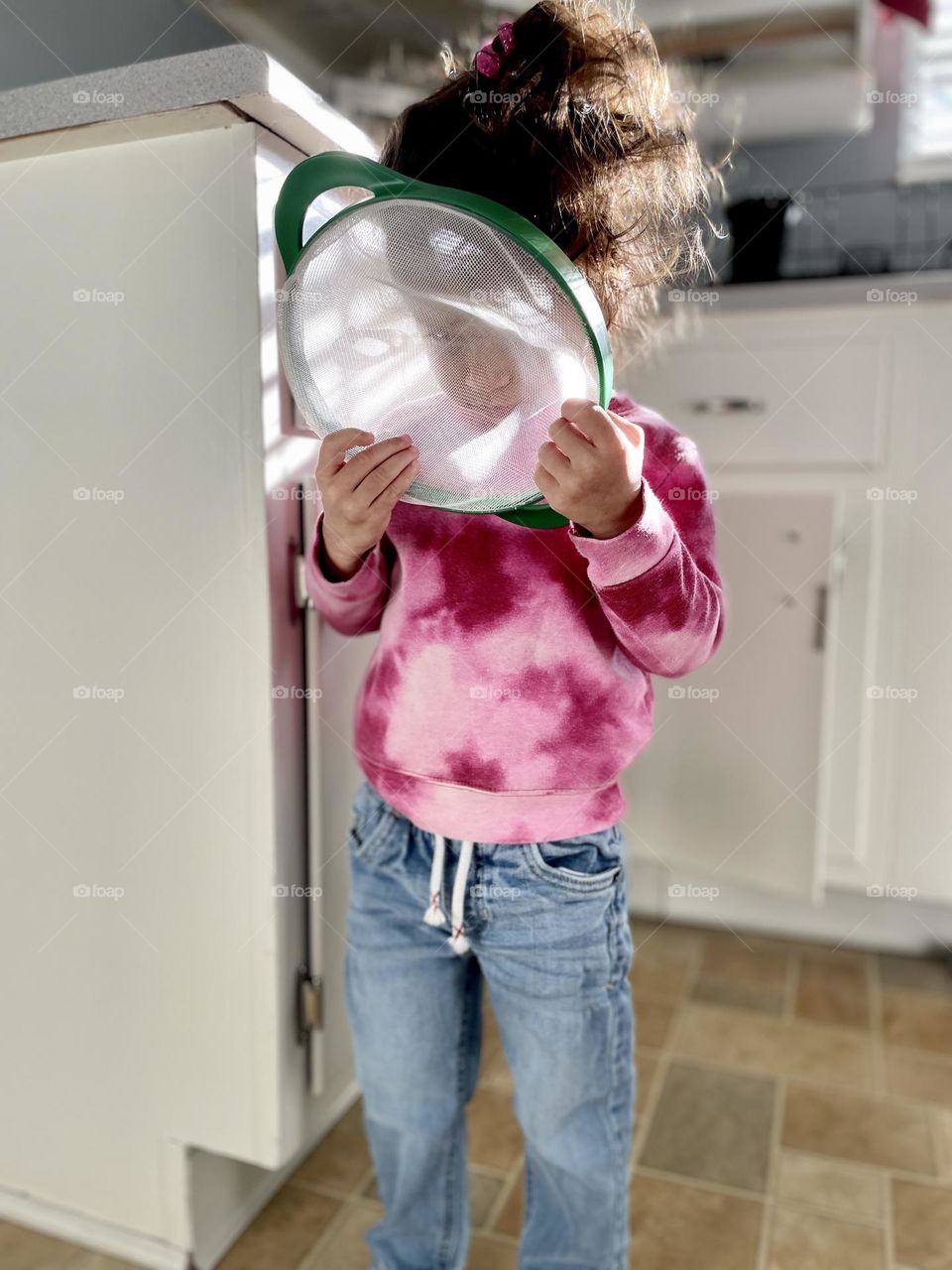 Little girl angrily looks through kitchen colander, little girl hiding in anger, toddler meltdown, toddler is angry, toddler girl shows angry emotion, toddler girl looks through colander in the kitchen, cute but angry little girl 