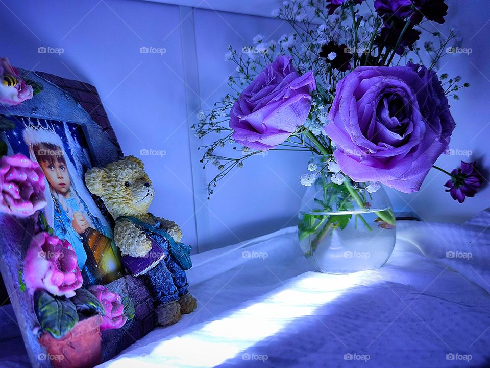 On the shelf is a jar of delicate purple roses, white small flowers and dark purple asters.  Nearby is a frame with a photo of a little girl. The ceramic frame is decorated with purple flowers and a bear in a purple vest. The shelf is illuminated.