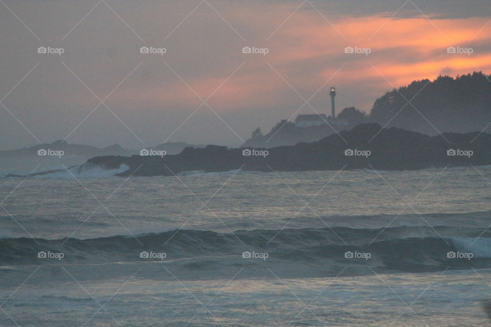 Sunset on the West Coast is always spectacular. This one with a view of the beach in the foreground, a rocky outlet with a lighthouse and its beam shining on the coast, and a multicoloured misty sky. 