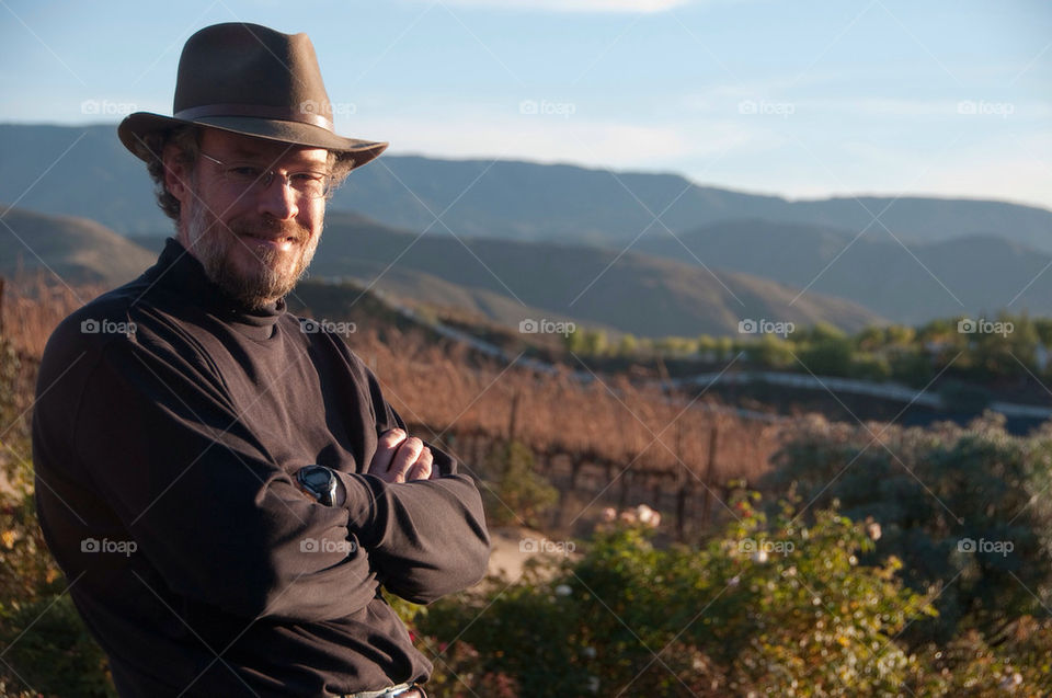 Man with a hat stands in front of the vineyard before the mountains