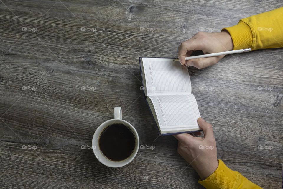 A man drinks coffee in the morning, works on the phone and makes notes in pencil in a notebook. Men's hands hold a white cup of coffee and a mobile phone in a yellow jacket. The hands lie on a gray wood background.