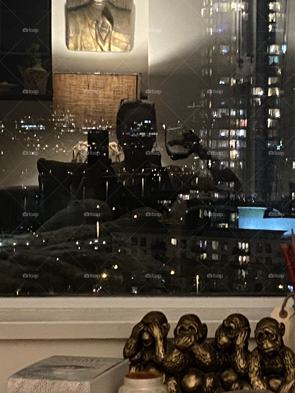 This is a reflection photo of a woman sitting inside drinking a glass of wine looking out onto the world the residential towers downtown living. Lamps are on in the background but you can see the residential towers and city life. 