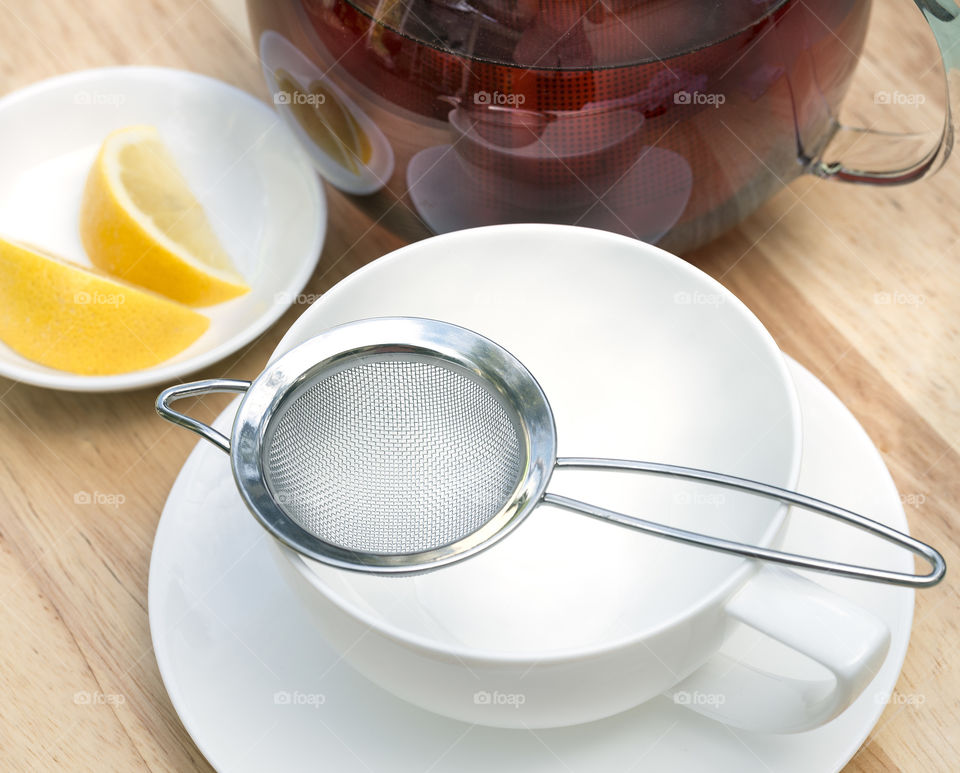 A white china cup and saucer with mesh tea strainer, teapot and lemon wedges on plate.