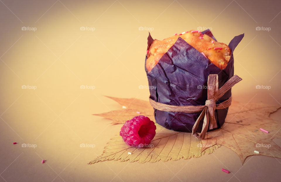 Autumn still life with pastries.  Muffin in brown baking paper with raspberries on autumn leaves with pink topping on a plain background.  Beginning of autumn