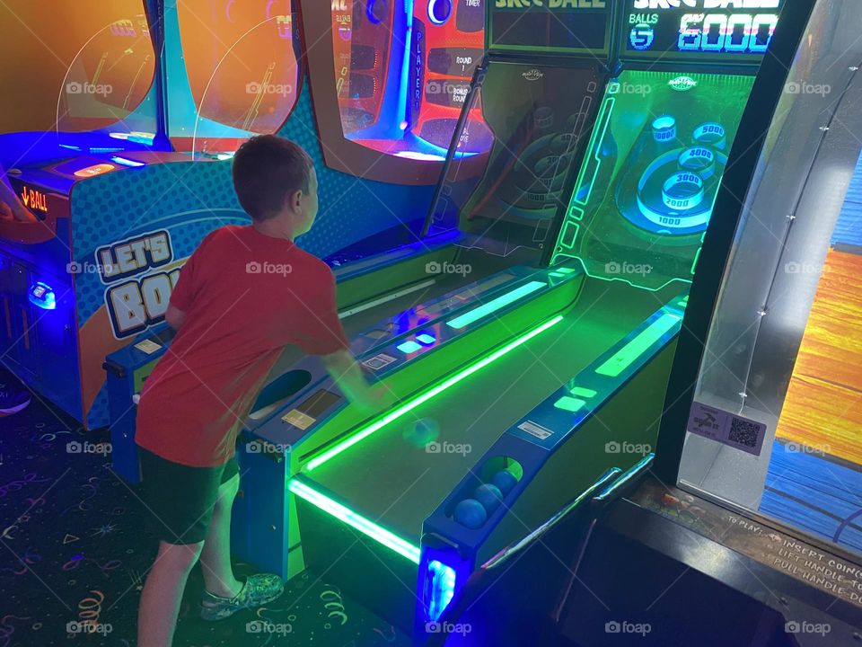 A young boy in a red shirt plays Skeeball in an indoor arcade. The arcade has bright neon green and blue lights. 