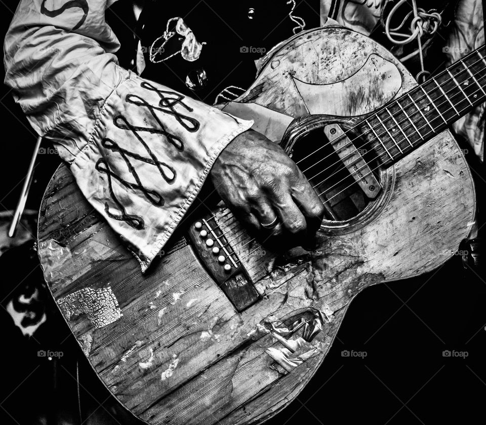 A hand in a bohemian shirt plays an old beat up acoustic guitar 