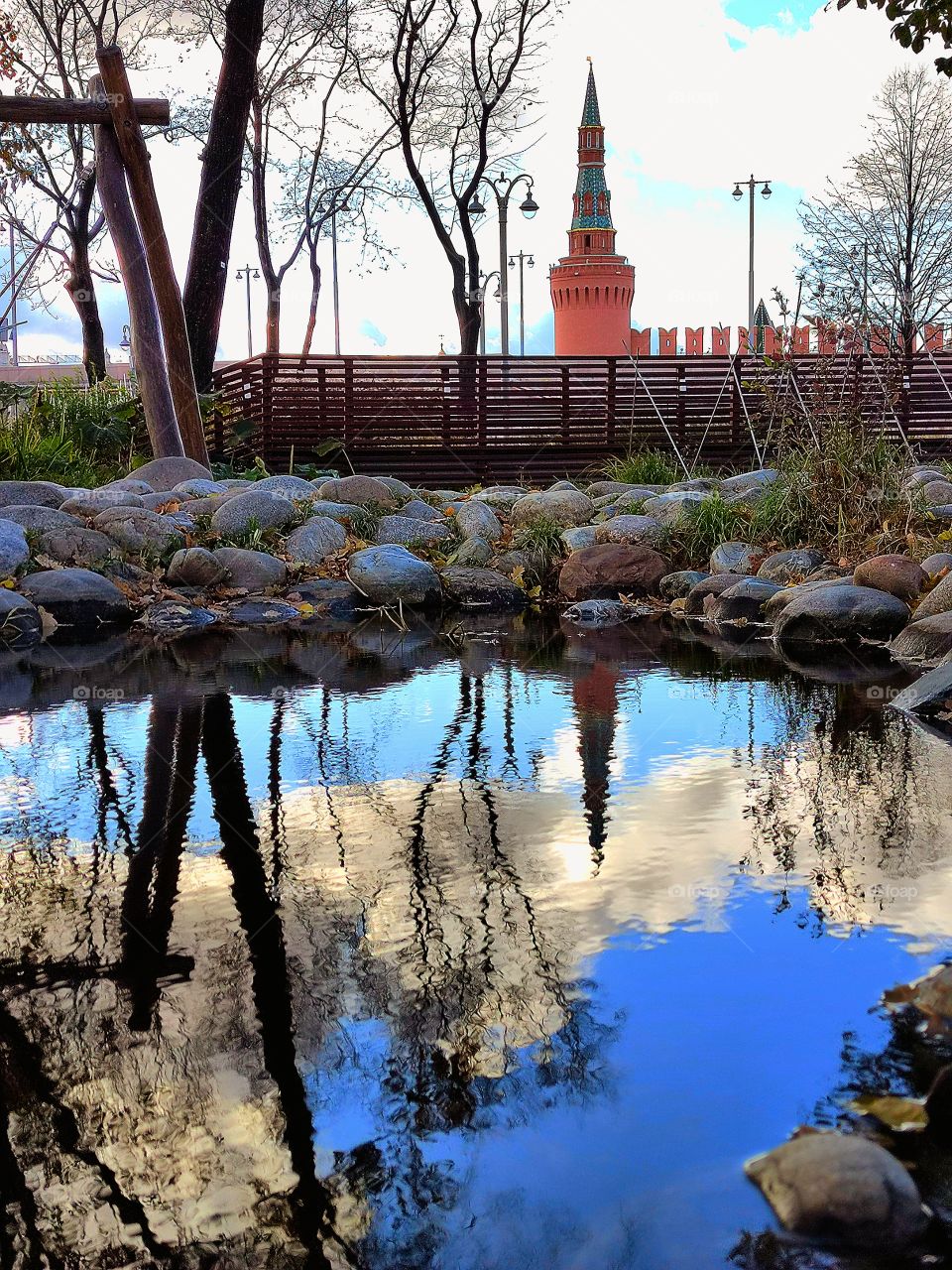 Reflection. The water of the pond reflects the Beklemishevskaya tower of the Moscow Kremlin, the blue sky with white clouds and trees. The pond is lined with stones on which fall leaves lie