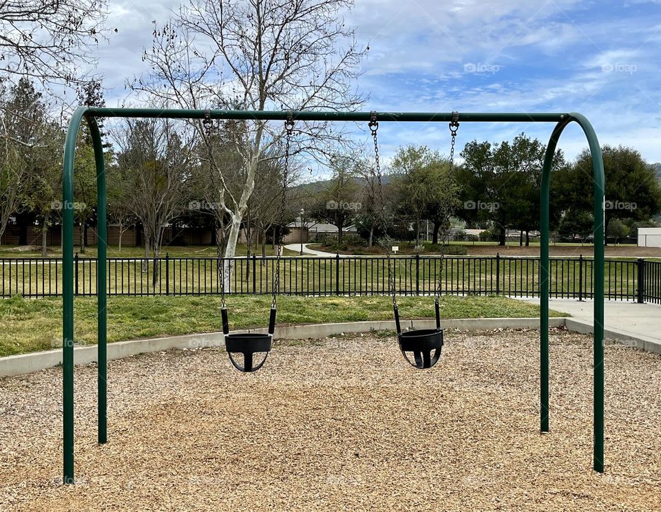 Classic playground equipment because all children love to swing!  Two toddler swings ready for playtime at the park with blue skies, green grass, and fenced in for safety