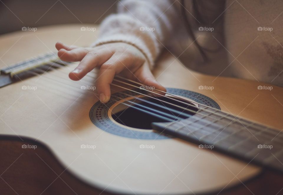 One hand of a little caucasian girl holds a guitar and strums
strings with fingers learning notes, side view close-up. Music education concept.