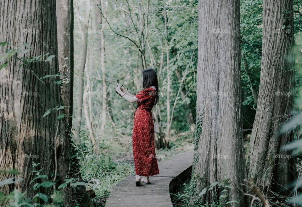 One young beautiful caucasian brunette girl with long flowing hair in a red dress takes a selfie on a smartphone, standing half-turn on a wooden winding path in the forest on a summer day, close-up side view.