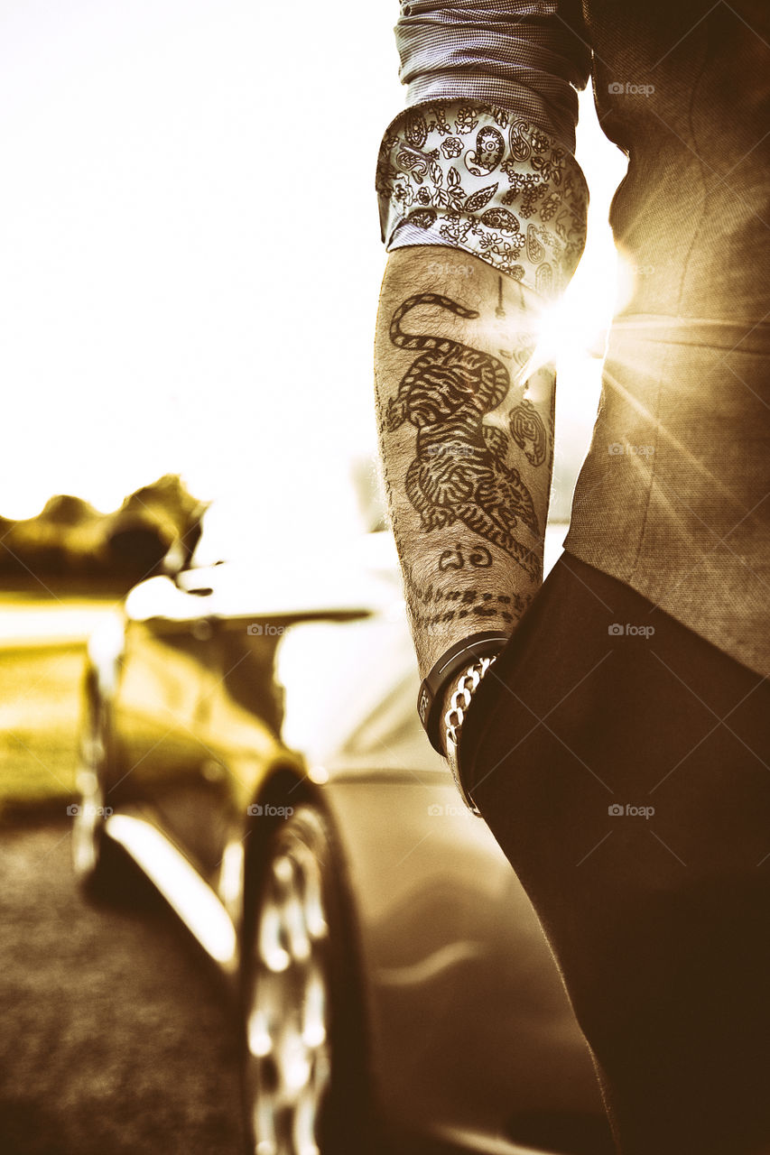Portrait of a young man with tattoo of tiger and Thai words on arm wearing smart casual business clothing standing beside a luxury sport car during sunny summer day