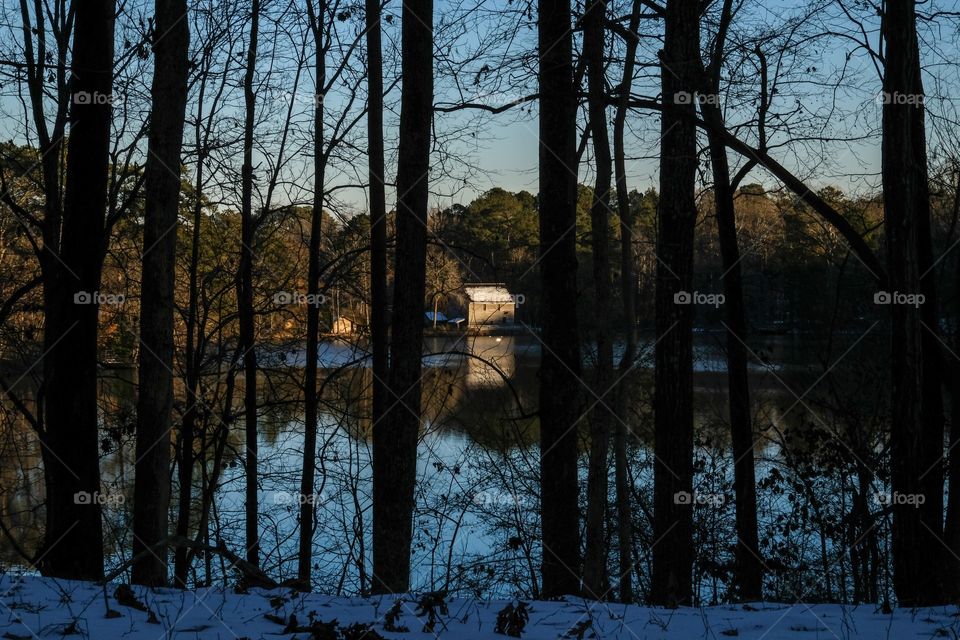 The last remnants of sunshine fall upon the old mill across the pond as seen through the trees, with a snow covered forest floor in the foreground at Yates Mill County Park in Raleigh North Carolina. 