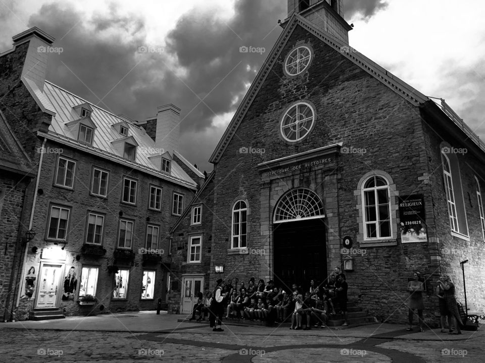 People sitting on the stairs of an ols church in french colonial cobblestone plaza of Quebec city during a cloudy evening