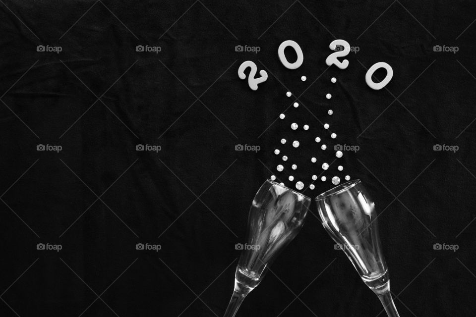 Flat lay of the numbers ‘2020’ above champagne glasses with rhinestone bubbles on black background in black and white - copy space on left side of landscape format