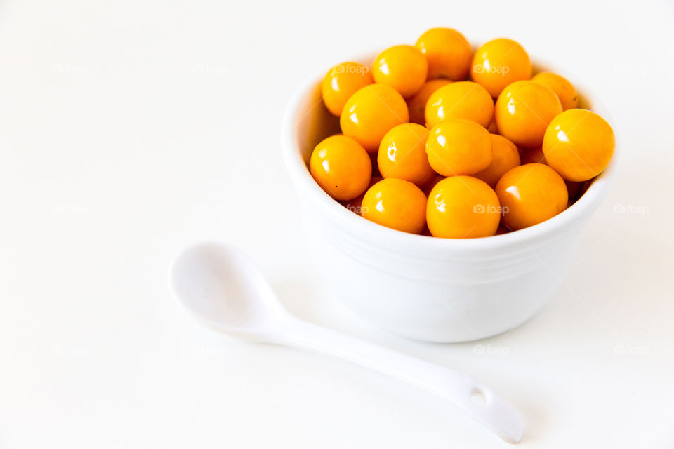 White bowl with fresh yellow gooseberries served with single white spoon on white background. Summer fresh healthy breakfast fruits.