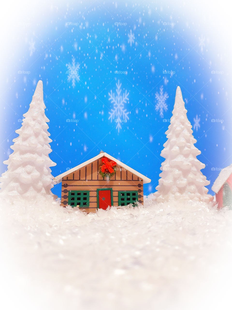 Winter miniature scene of a log cabin and two snow covered pine trees surrounded by snow with a blue sky and snowflakes.