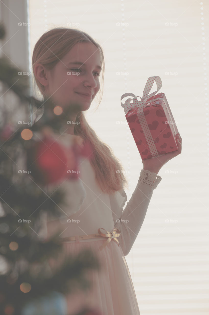 Happy smiling girl wearing pink dress standing behind a tree and  unpacking Christmas gift