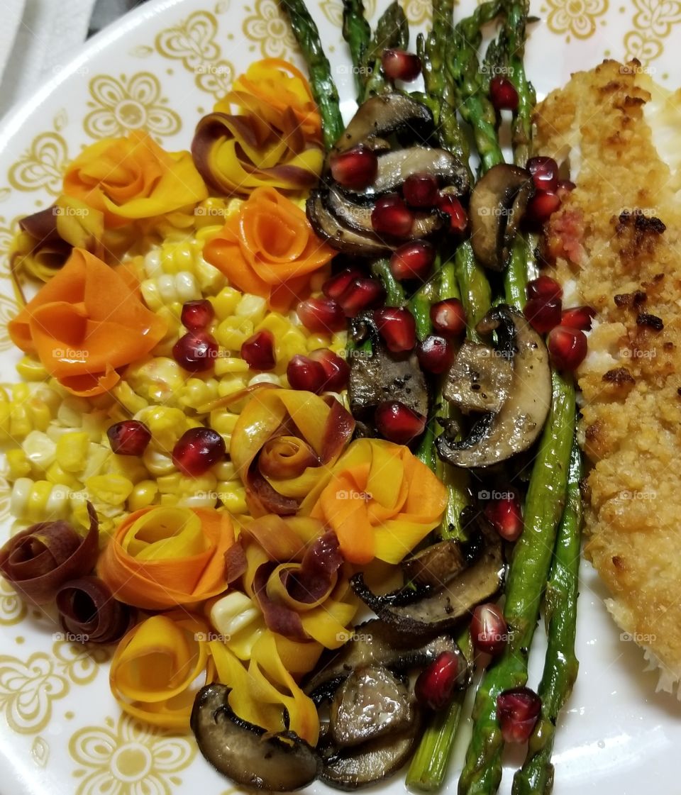 Seasonal Autumn harvest dinner of baked haddock with the last corn of the season, oven roasted asparagus and mushrooms. Thin slices of purple, white and orange carrots were steamed, then folded into roses. The plate was sprinkled with fresh pomegranate arils for an added accent of color, texture, and a sweet-tart burst of flavor.