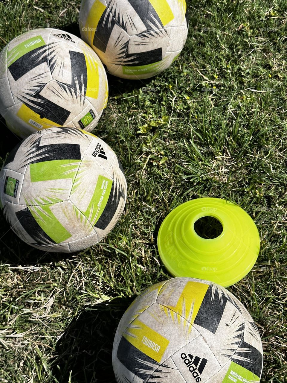 Four Adidas Tsubasa training soccer balls with green and yellow design with fluorescent yellow/green training cone at a practice on grassy soccer field