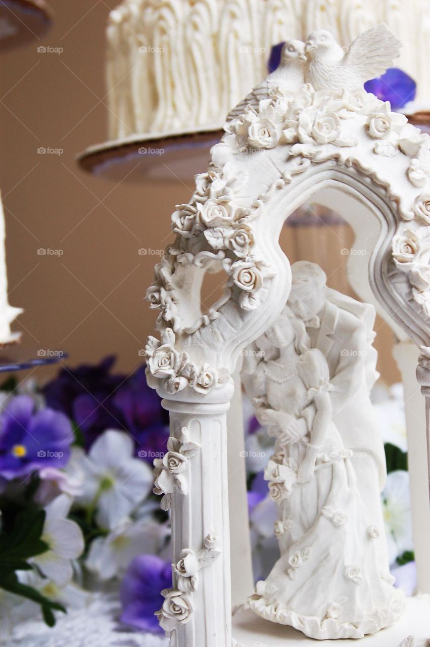 white wedding cake topper couple in loving embrace under arch of roses on a table with purple flowers