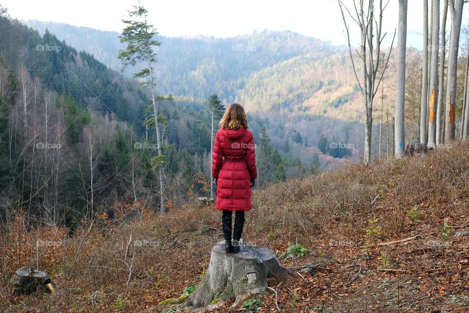 A woman in red coat standing on a stump with her back to the viewer and watching at the forest and mountains at Karlovy Vary neighborhood.