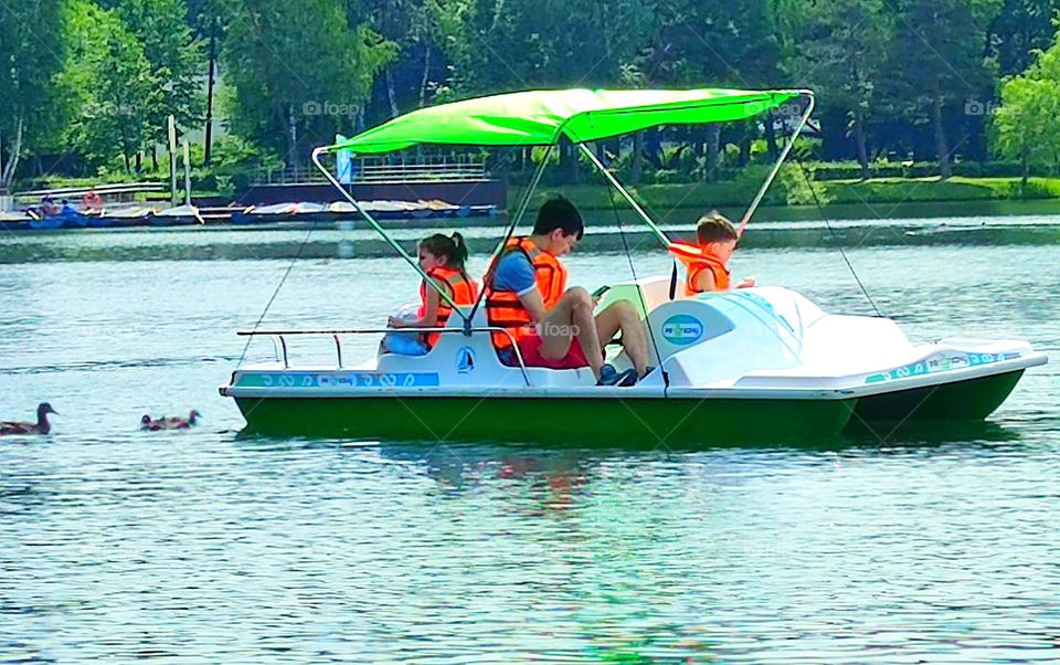 Summer in the city.  Pond in the park.  A catamaran with a sun-protective green roof floats on the water.  Father, son and daughter are sitting in the catamaran.  Ducks follow the catamaran