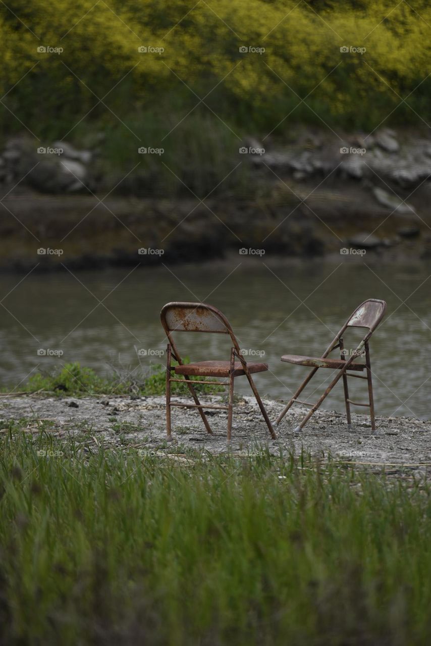 Left behind, two metal chairs rusting, abandoned by fishermen along a river.