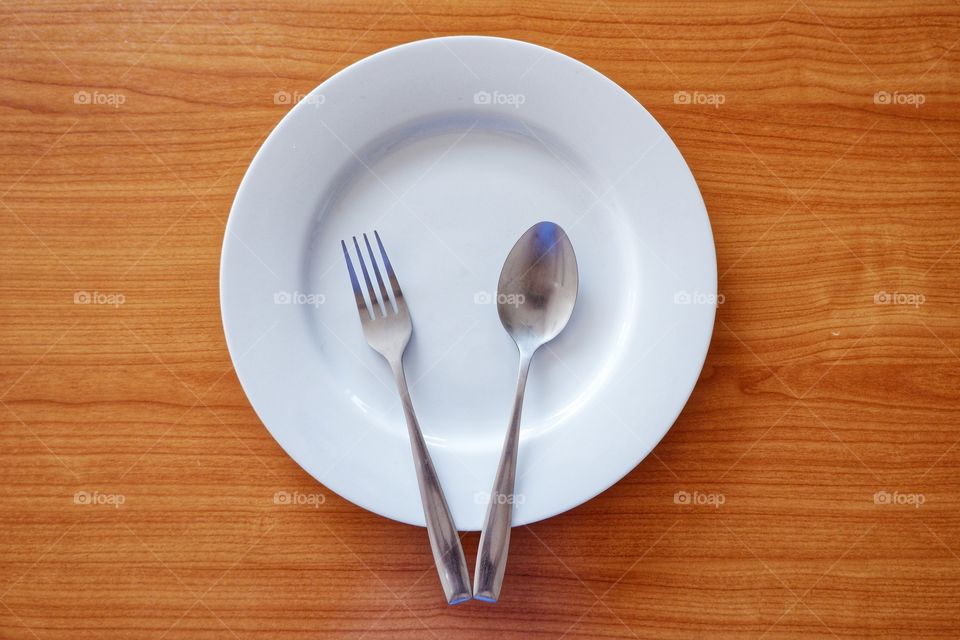 Empty white plate with spoon and fork on blown wooden background.