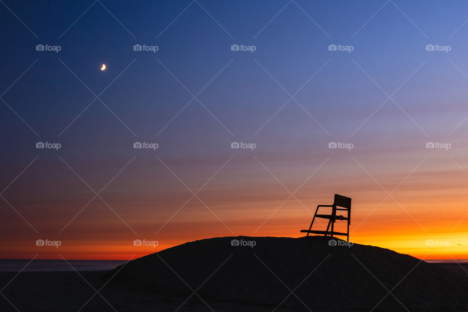 Silhouette of a lifeguard chair on top of a sand tower on the beach. Crescent moon and sunset sky in the background