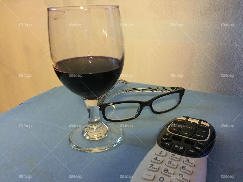 A glass of red wine a pair of Of reading glasses and a television remote control 