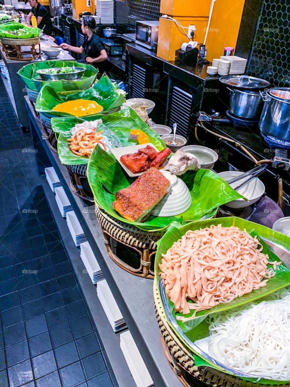 a long table they arrange trays and display food on it , we can also see the bowls, plates and pots. waiters sitting there, each person will be in charge of a few different items , the foods is placed on a banana leaf