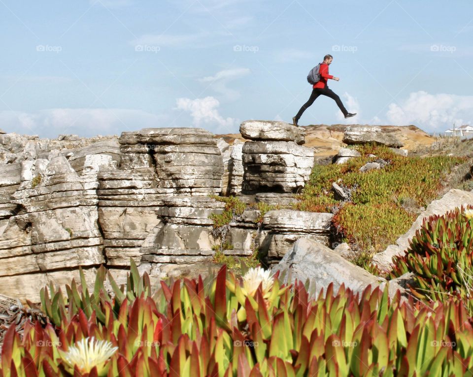 A man leaps over a gap in the rocks, with blue sky behind him and colourful plants in the foreground 