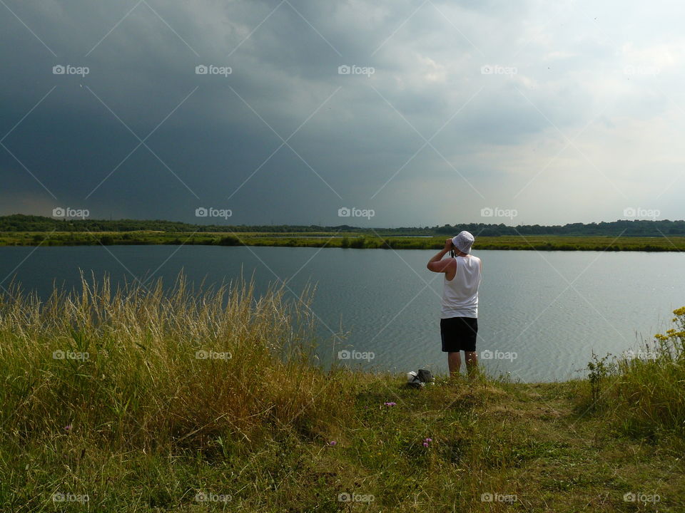 Man Holding Binoculars Looking Into Stormy Skies Standing By A Beautiful Large Lake