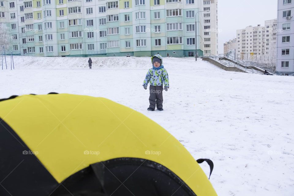 A small, carefree boy walks in the white snow in winter and rides a tubing in the park, near trees in the snow.