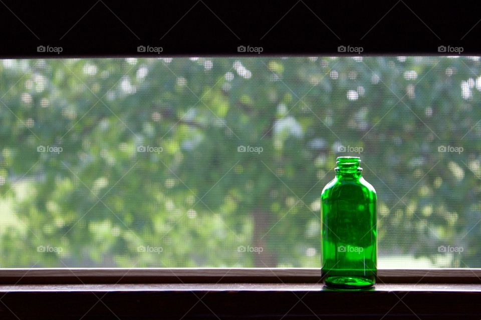 A small, empty green bottle on a windowsill by an open, screened window and a dark brown shade partially pulled down with a blurred tree visible in the background 